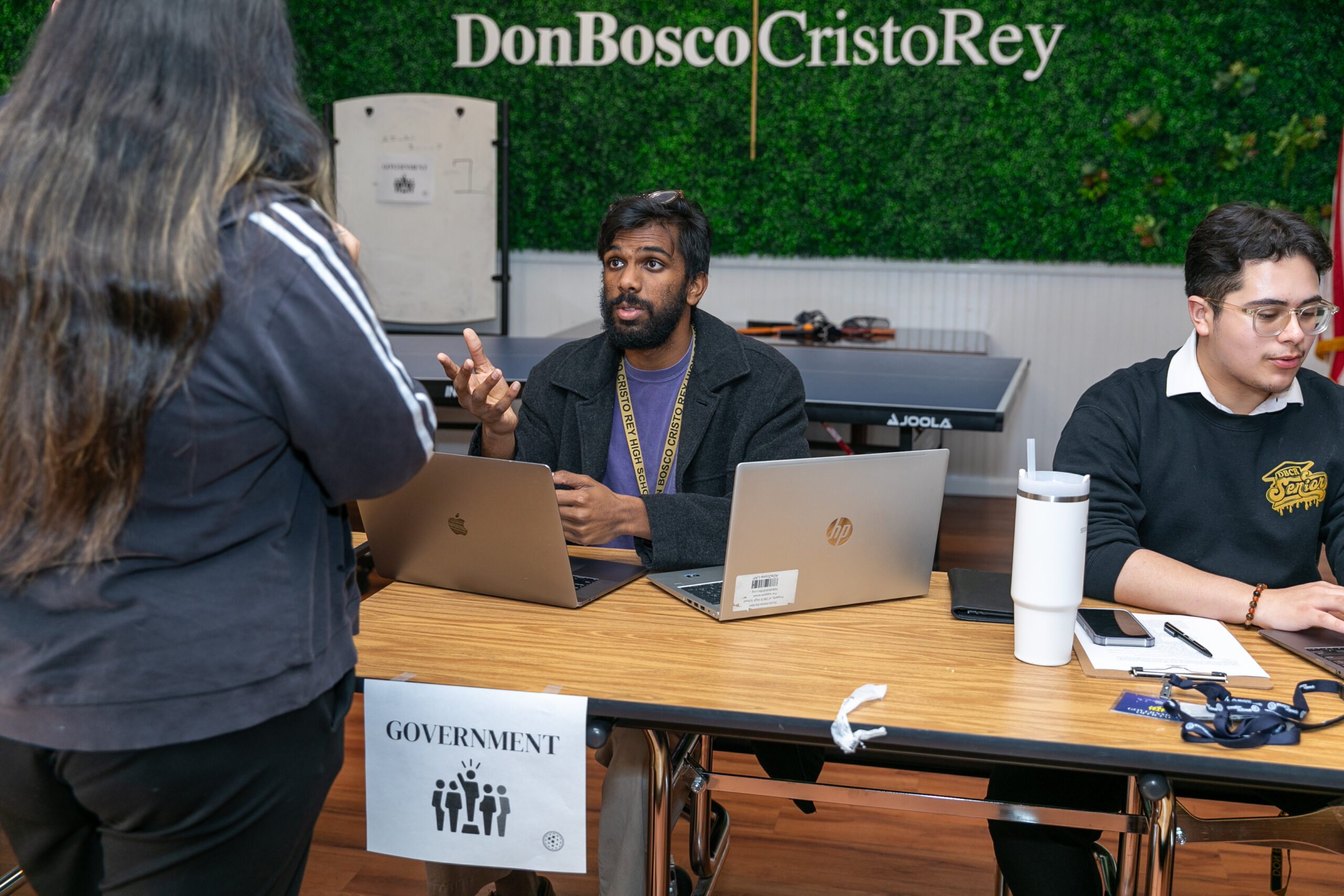Two people sit at a table with laptops under a “Don Bosco Cristo Rey” sign, discussing operation outbreak with a person standing in front of them. A “Government” sign is taped to the table.