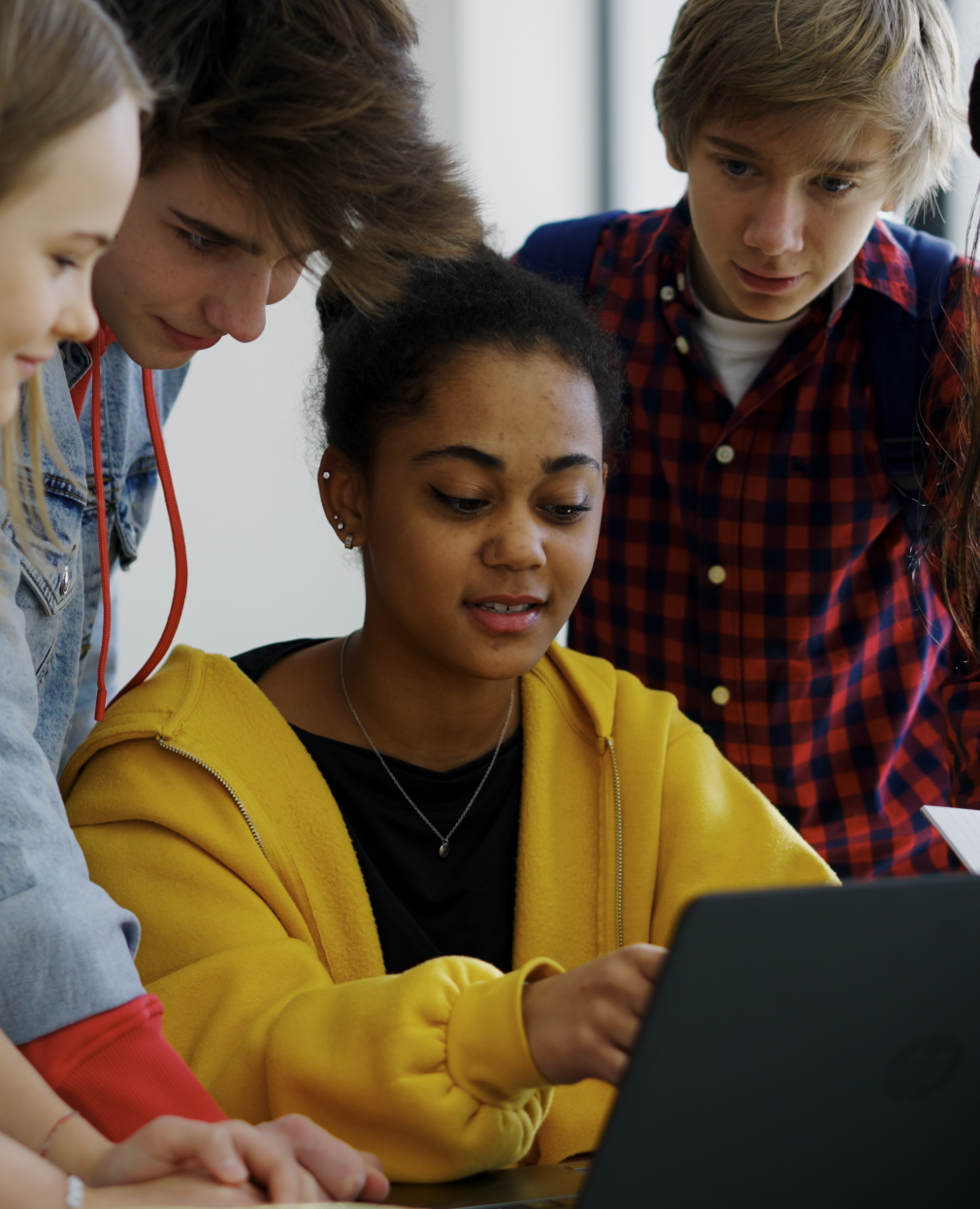 A group of teenagers gathers around a laptop, with one girl in a yellow hoodie pointing at the screen while the others watch attentively and take notes.