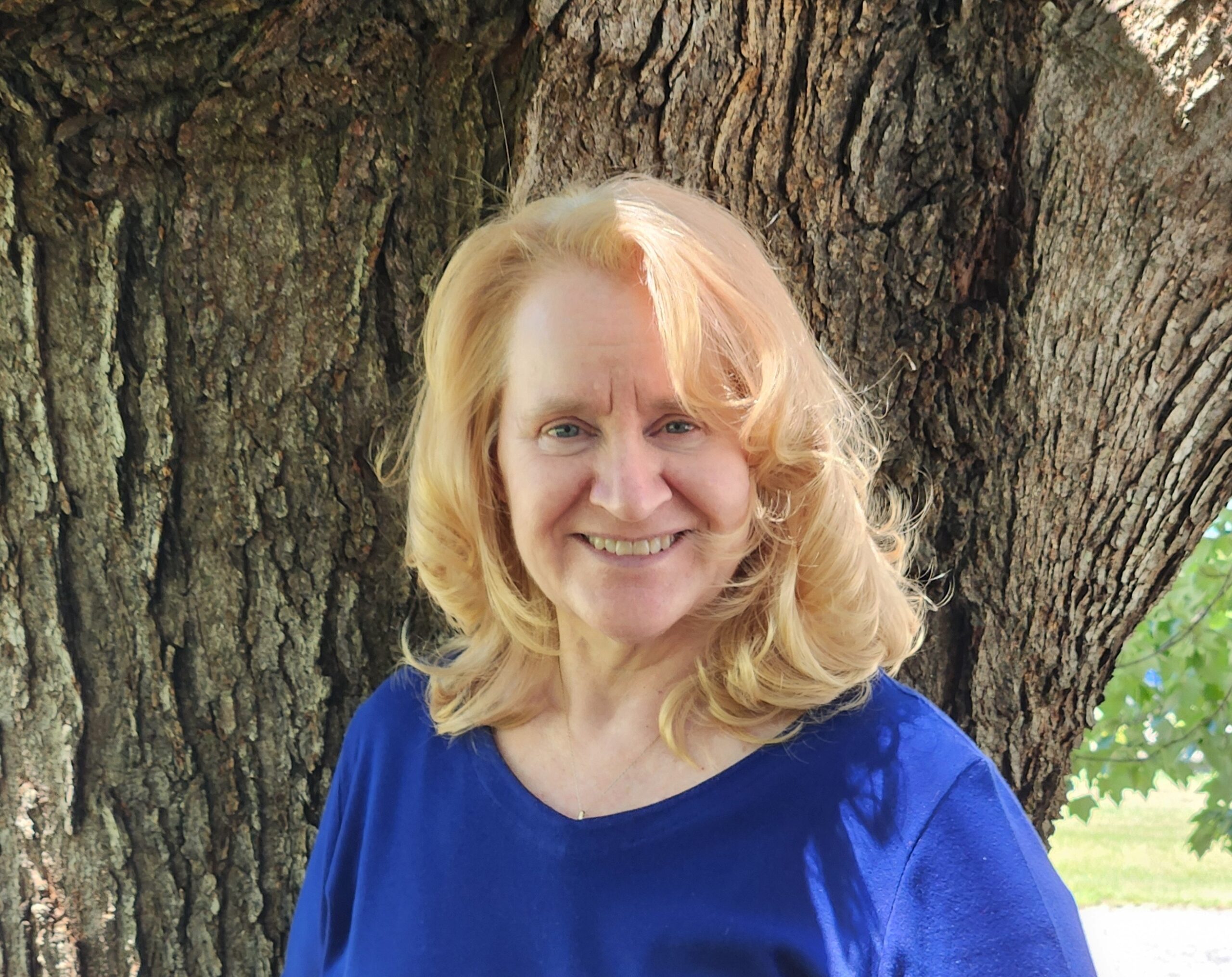 A woman with wavy blonde hair wearing a blue top smiles while standing in front of a large tree trunk outdoors on a sunny day.
