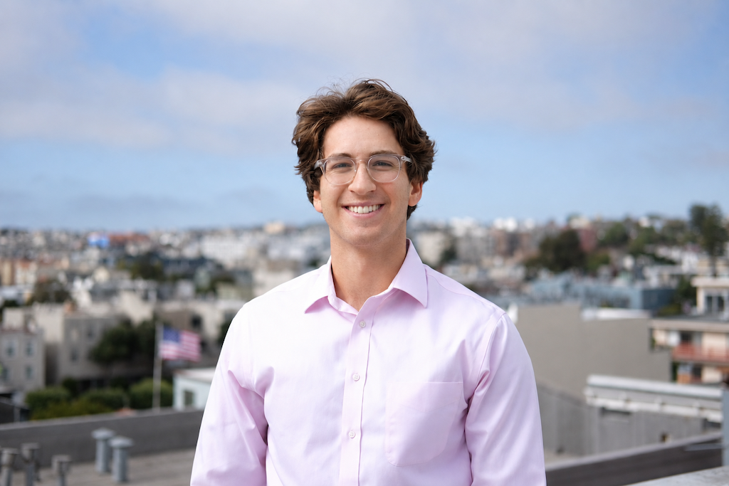 A young man with glasses and brown hair, wearing a light pink shirt, smiles while standing outdoors with a cityscape and an American flag blurred in the background.