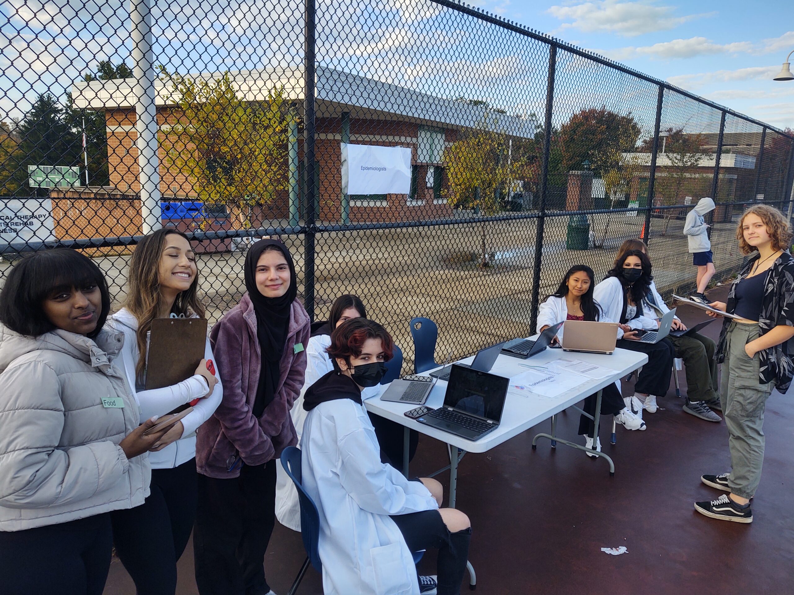 A group of young people stand and sit by a table with laptops and papers outside on a basketball court. Some are smiling, and a few wear white coats or masks. It appears to be an outdoor event or activity.