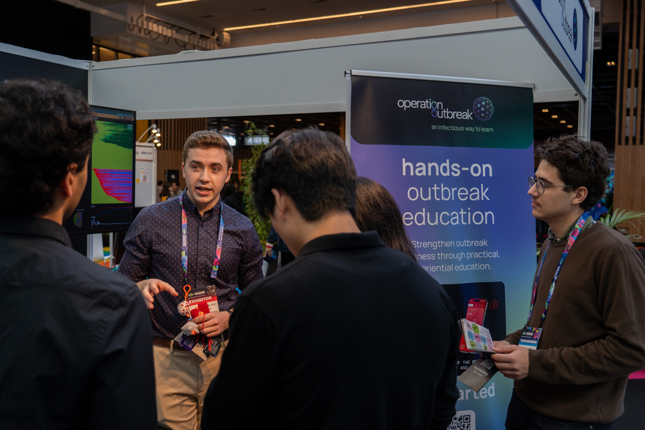 A man speaks to a small group at an expo booth with a “hands-on outbreak education” banner and a computer screen; attendees listen and hold event lanyards.