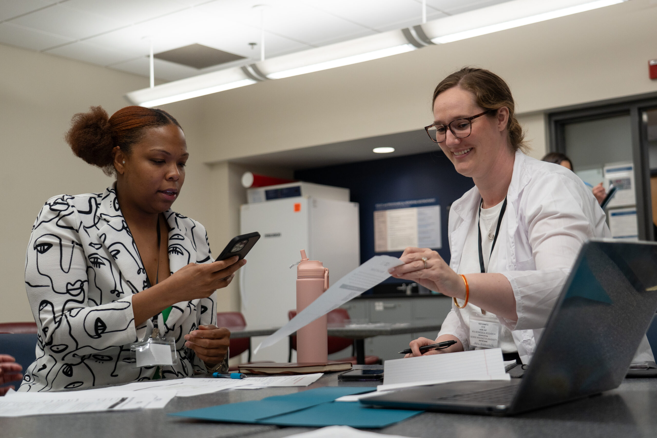 Two women sit at a table with papers, a laptop, and a water bottle. One woman smiles while handing over a document; the other looks at her phone. They appear to be working together in an office or classroom.