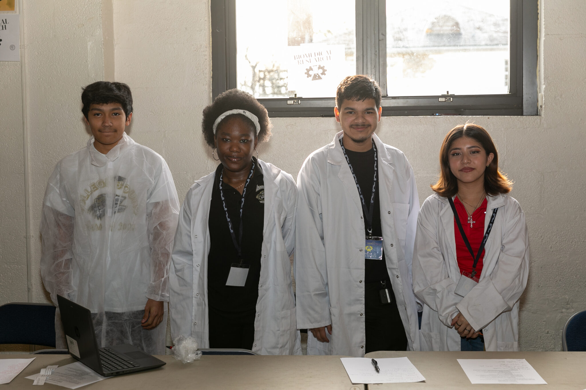 Four students wearing lab coats stand side by side in a classroom, with a window behind them and papers and a laptop on the table in front.