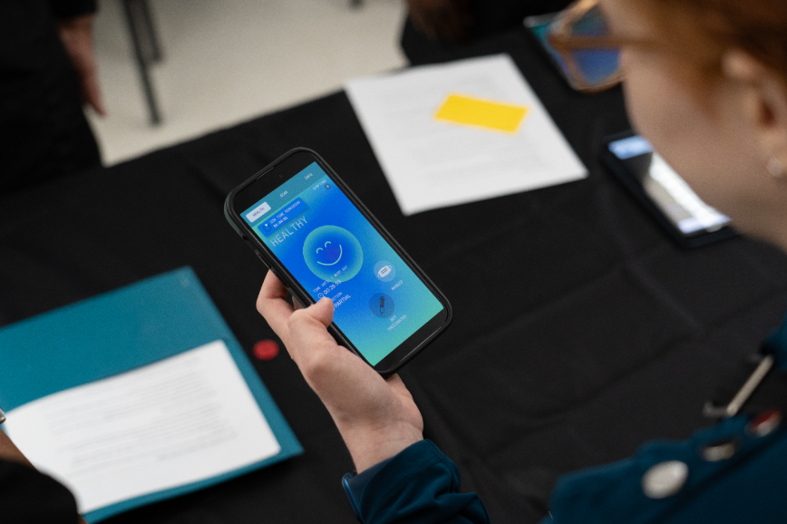 A person holds a smartphone displaying a health app with a smiling face. Papers and folders are on a black table in the background.