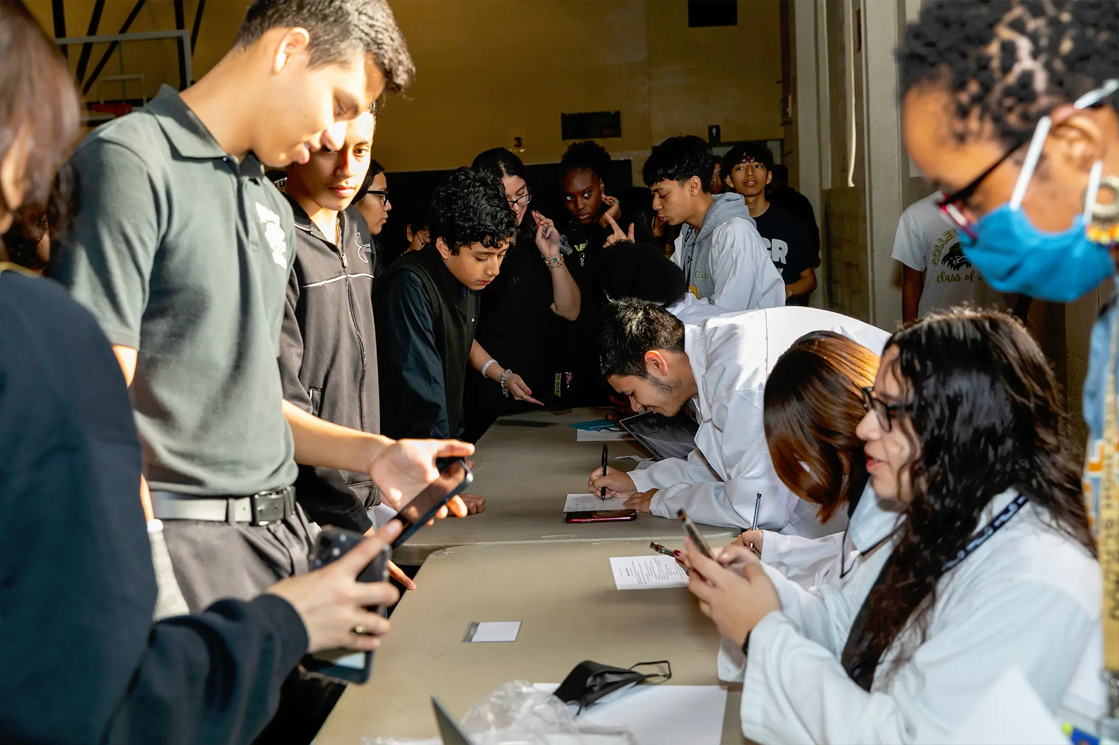 A group of people stands in line at a table while others, some in white coats, assist them during a simulation