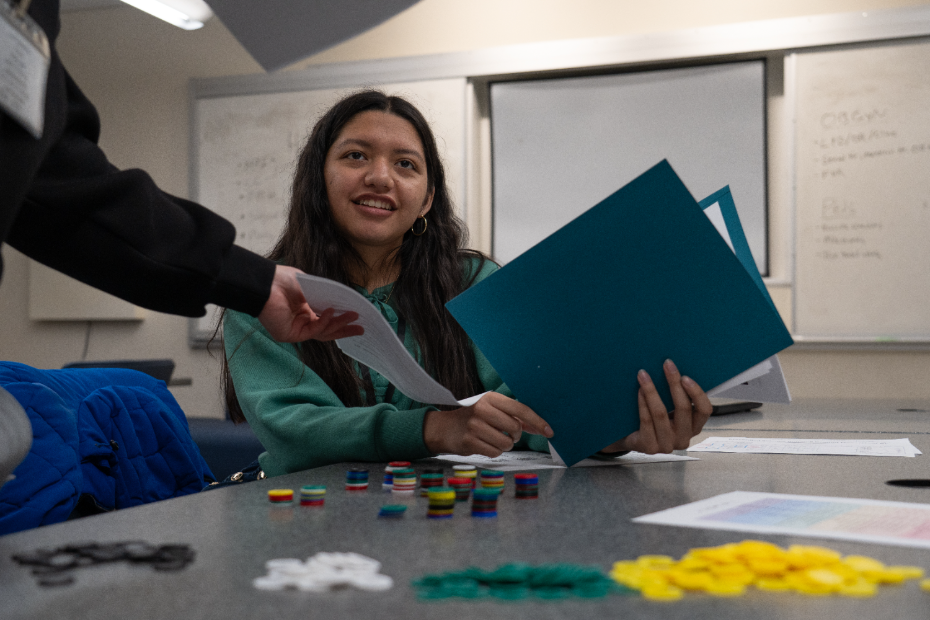 A person sits at a table holding folders and smiling, with colorful game pieces and papers spread out in front of them. Another person hands over a sheet of paper.