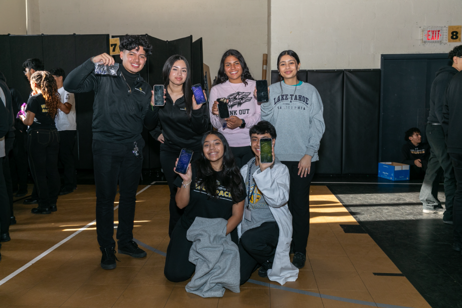 Six smiling students stand indoors, holding up phones displaying screens. They wear casual and school-branded clothes. Other people and a blue box are visible in the background on a gymnasium floor.