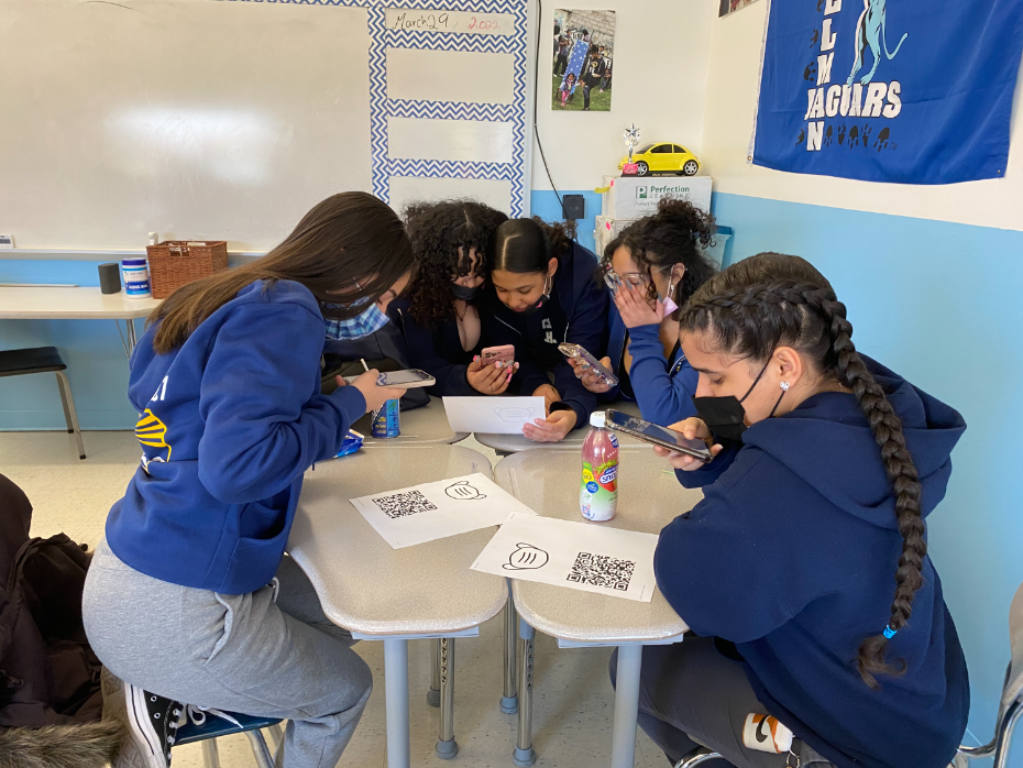 Five students in a classroom sit around a table scanning QR codes with their phones, focused on the activity. They wear blue uniforms and the room has school banners and colorful decor on the walls.