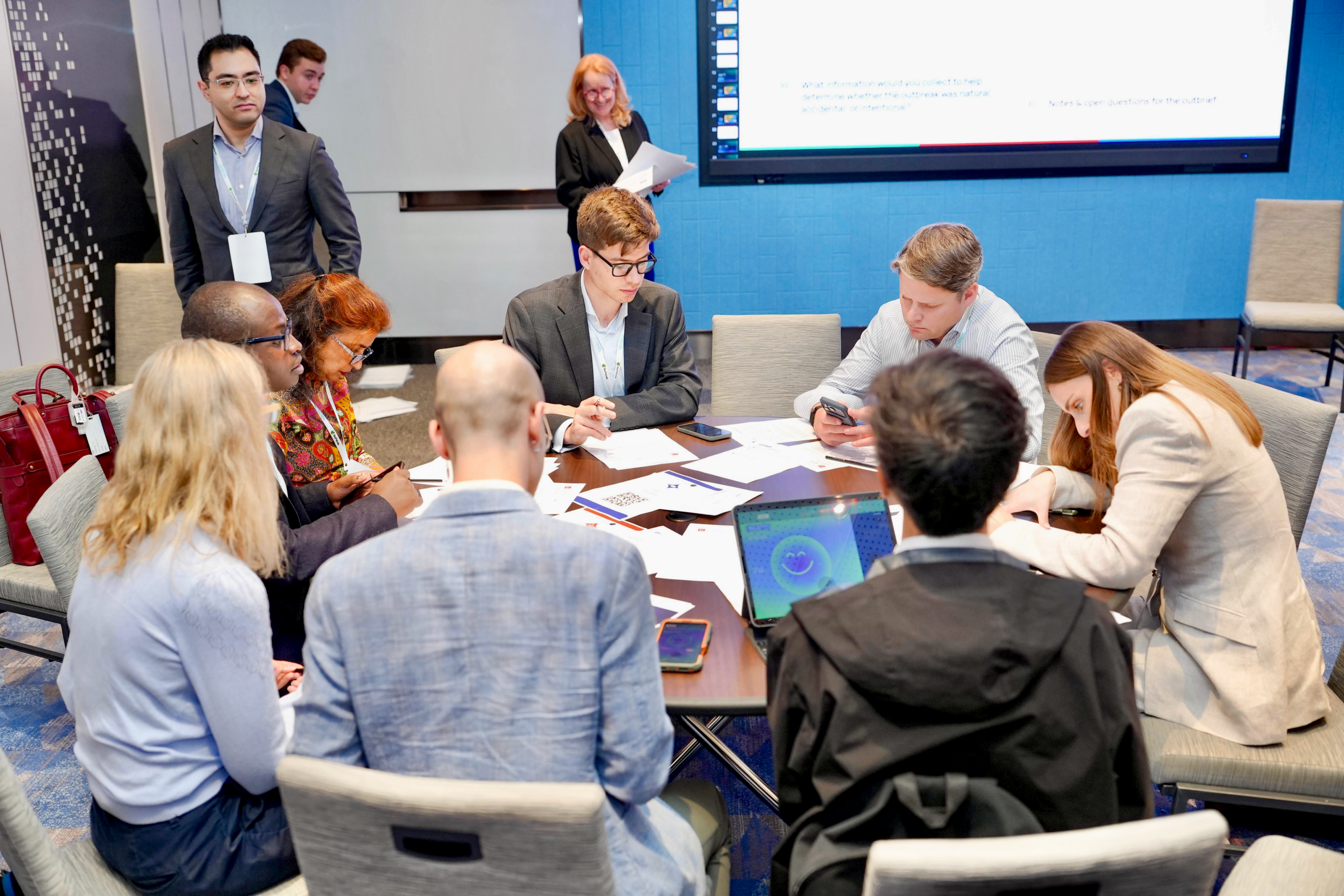 Participants around a table during a tabletop exercise, working through printed injects and QR sheets on their phones while a facilitator presents the discussion question behind them