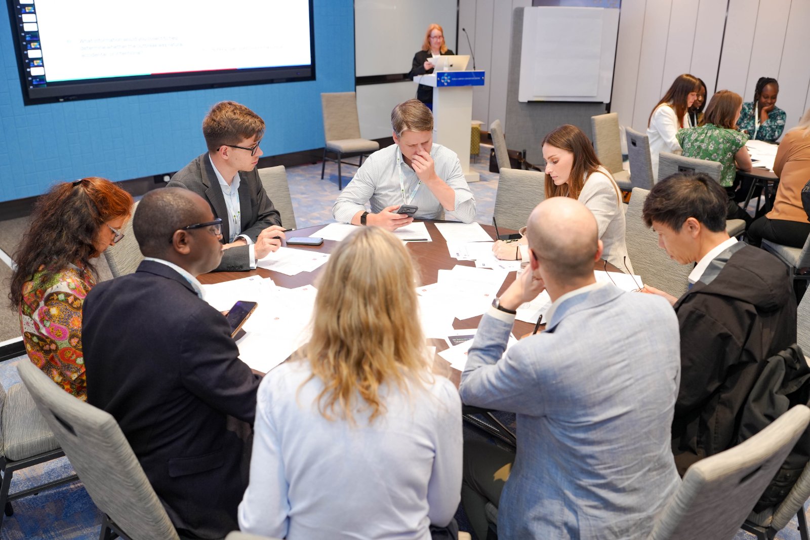 Two tables of participants at the Kuala Lumpur exercise, working through printed injects while a facilitator presents from the podium