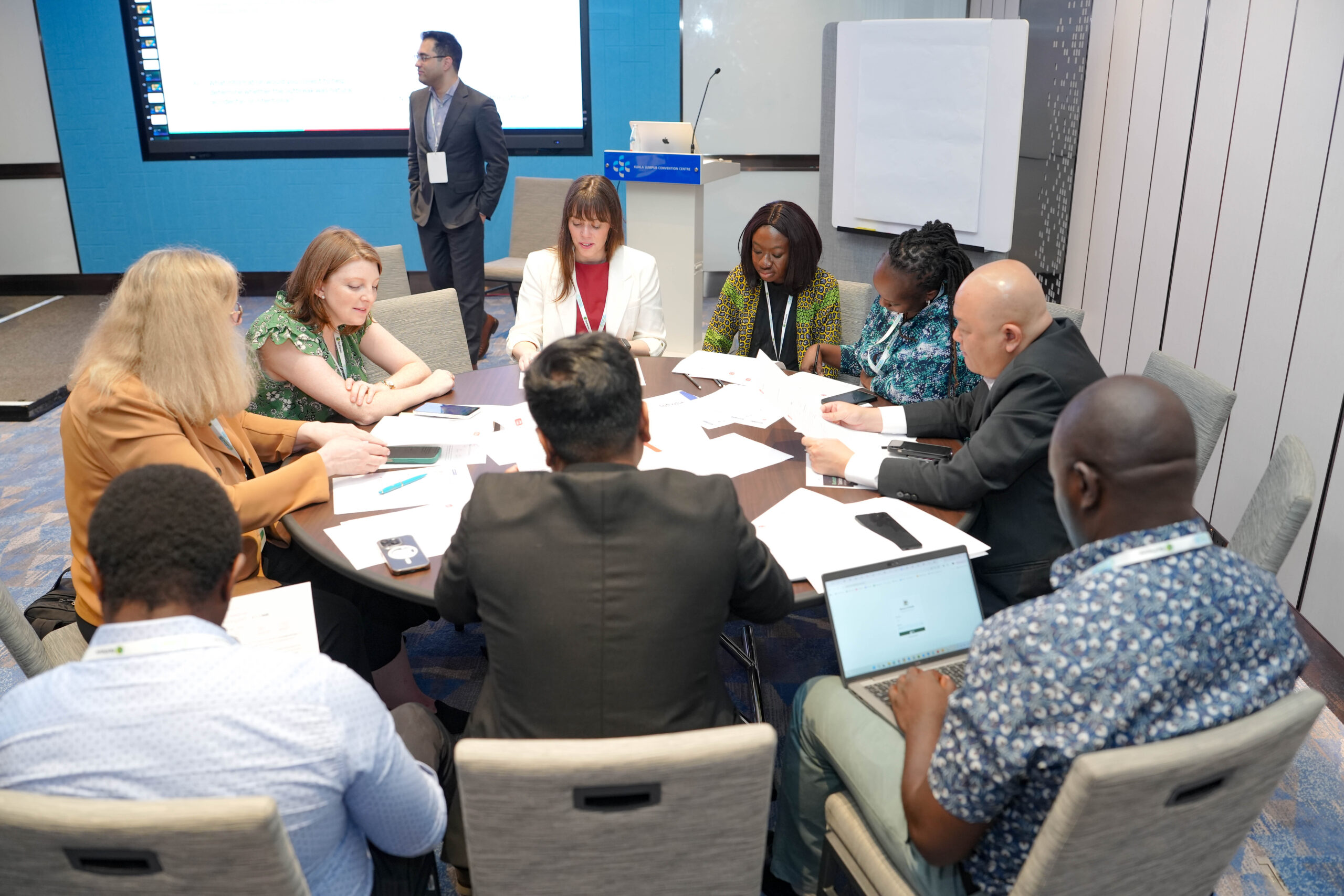 Participants at a tabletop exercise working around a table with phones, printed injects, and countermeasure QR sheets while a facilitator presents a discussion question