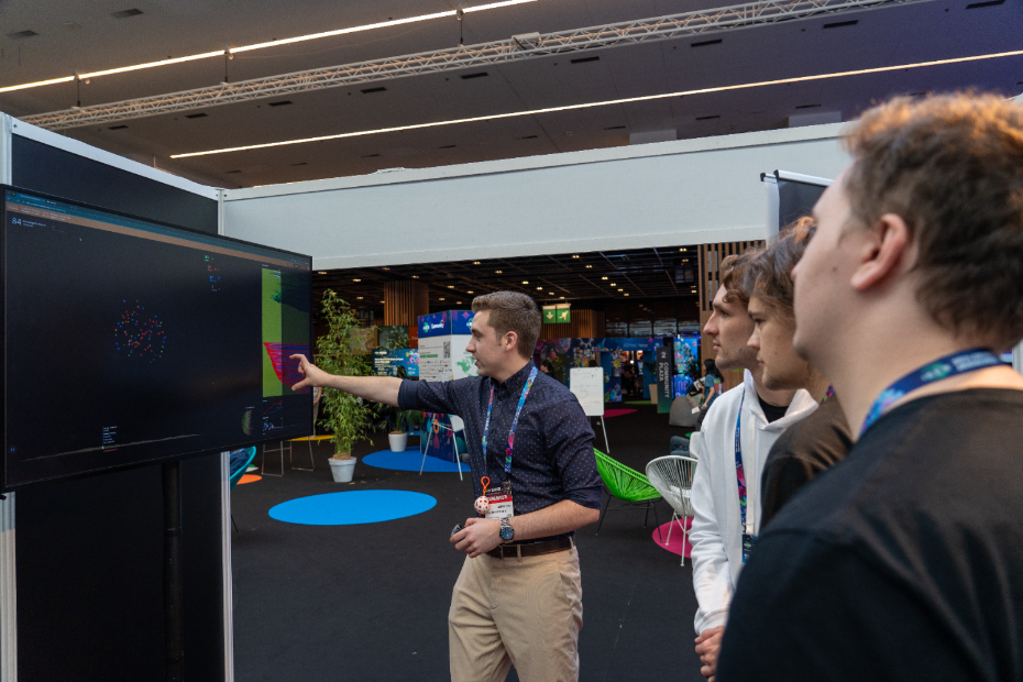 A man points at a large computer screen while three people watch attentively at a tech event in a modern, well-lit venue.